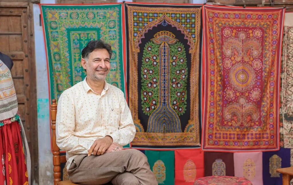 A wide shot of a traditional sunlit artisan studio in Kutch, Gujarat, with white walls adorned with finished Rogan wall hangings of the "Tree of Life" and sacred deity portraits, showing the tools of the craft like the metal tulika and ceramic pots of pigment.