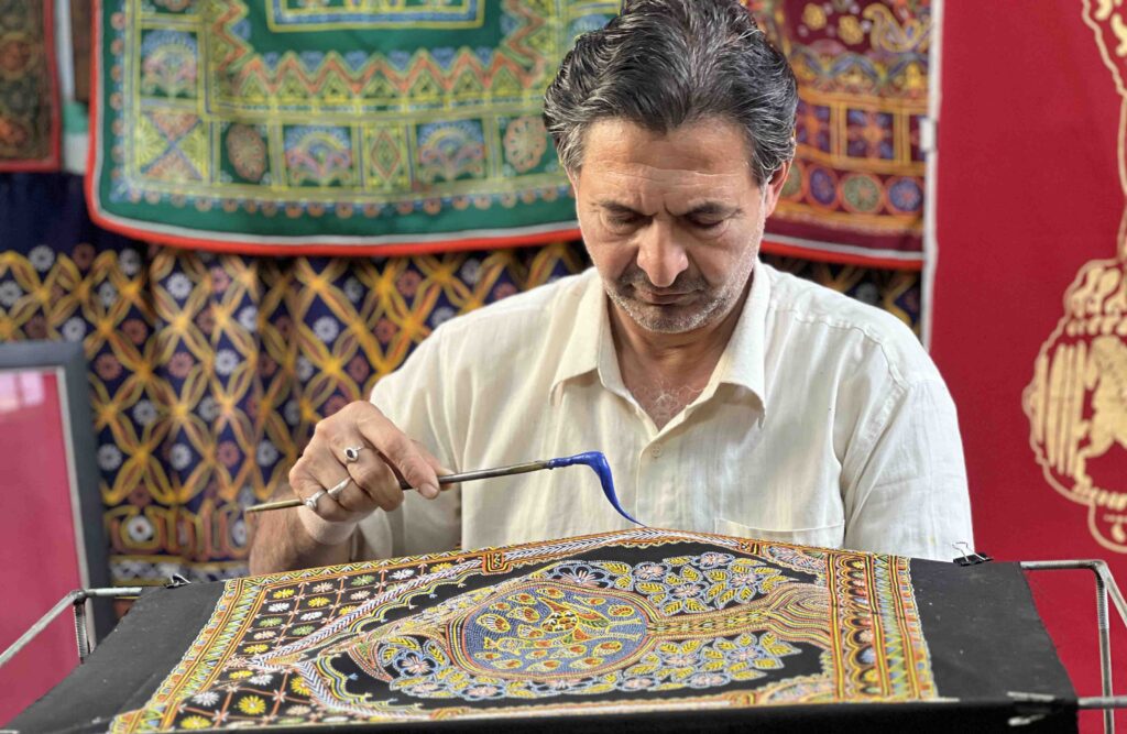Master artisan Ashish Shantilal Kansara demonstrating the "liquid thread" technique by pulling a strand of blue Rogan paint with a metal stylus over a dark fabric.