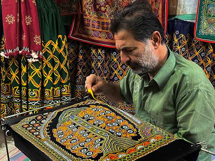 Master artisan Ashish Kansara performing authentic Rogan Art, using a metal stylus to paint a detailed Hindu deity with castor oil paste on dark fabric.
